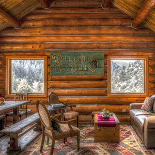 Cozy cabin interior with rustic furniture and "Cabin Welcome - Wood & Metal Wall Art" mounted on log wall above windows.